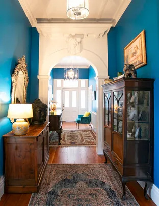 A bright hallway with blue walls, wooden furniture, decorative mirror, and a glass cabinet, featuring ornate details and rugs.