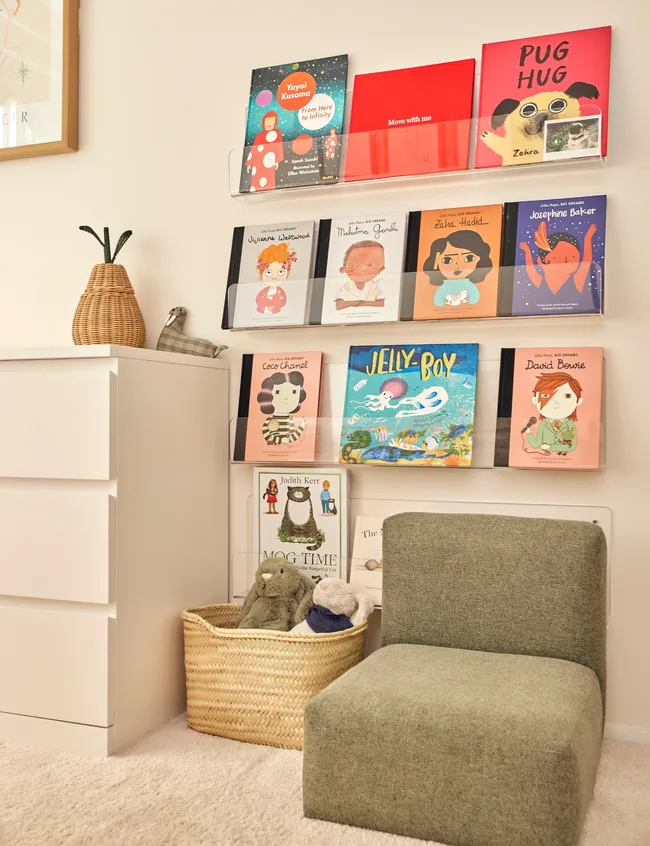 Children&rsquo;s room with a green chair, books on wall shelves, wicker basket with plush toys, and a potted plant on a dresser.