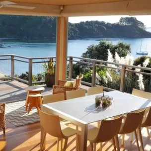Outdoor dining area with a white table and chairs overlooking a scenic bay and lush greenery.