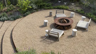 Gravel patio with circular fire pit, surrounded by white wooden chairs and greenery.