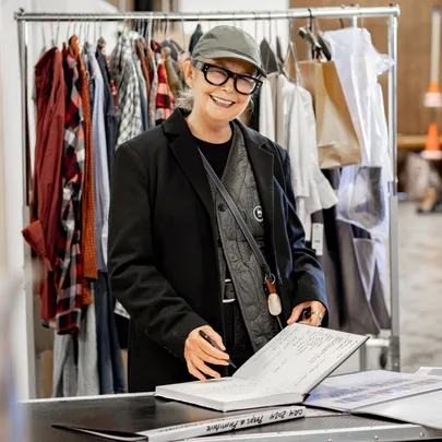 A person wearing glasses and a cap smiling, standing by a rack of clothes and writing in a large notebook.