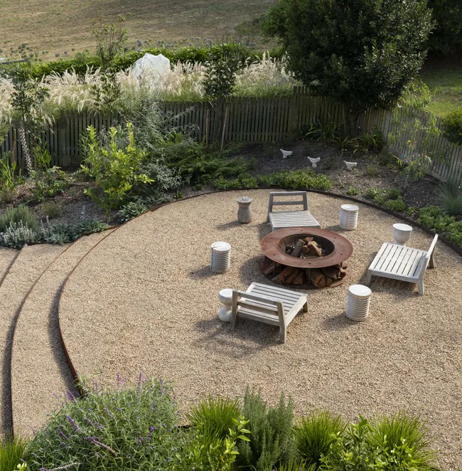 Outdoor fire pit with white wooden chairs and stools on pebbled ground, surrounded by greenery and a wooden fence.