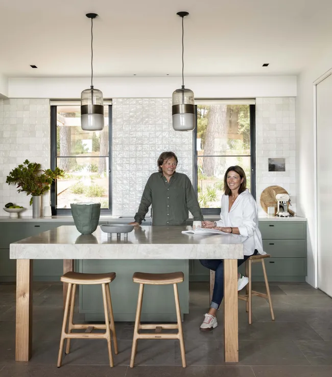 A man and woman in a modern kitchen with green cabinetry, light wood stools, and large pendant lights.