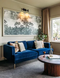 Blue sofa with cushions, large landscape artwork, wooden coffee table, glass bottle, book, and window with curtain.