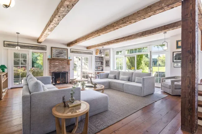 Spacious living room with exposed wooden beams, gray sofas, a brick fireplace, large windows, and wooden floors.