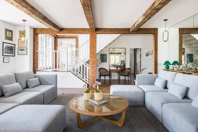 Spacious living room with gray sofas, wooden beams, round coffee table, and staircase leading to dining area.