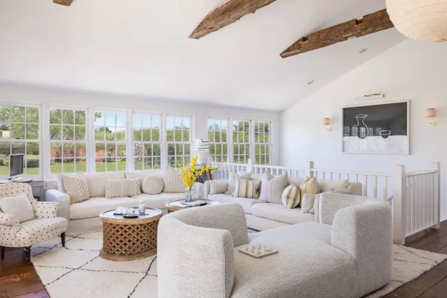 Bright living room with white sofas, wooden beams, large windows, a round coffee table, and artwork on the wall.