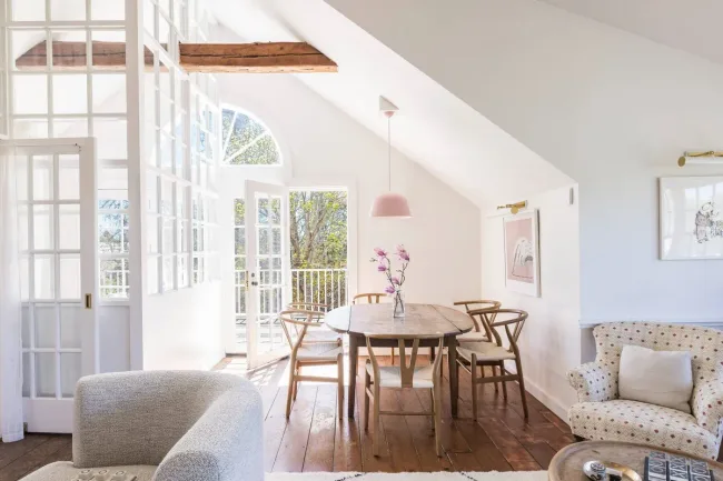 Bright room with a wooden dining table, chairs, pink pendant light, and large windows opening to a balcony.