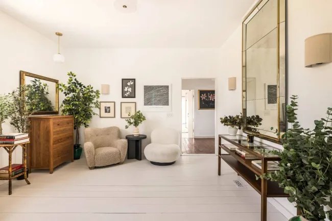 Bright living room with armchairs, plants, wall art, large mirror, and wooden furniture on white flooring.