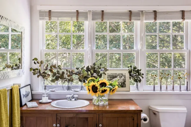 Bathroom with sunflowers on the wooden counter, large windows, plants, and yellow towels by the mirror.