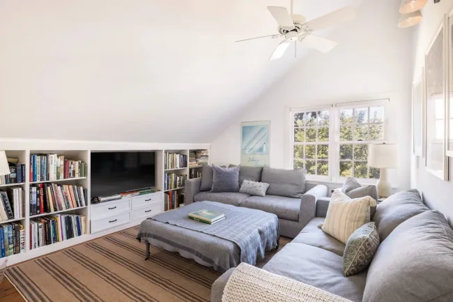 Bright room with a gray sectional sofa, bookshelves, TV, ceiling fan, and large window with trees outside.