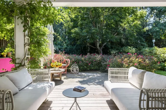 Patio with white sofas, a round table with sunflowers, and lush greenery surrounding a wooden deck.