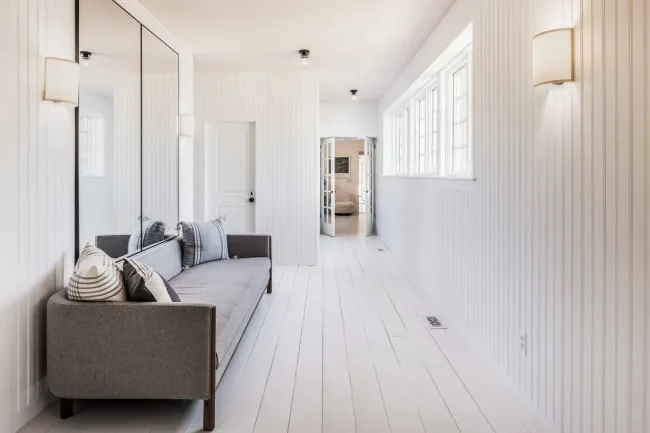 Bright hallway with a grey sofa, striped and plaid pillows, white wood paneled walls, large mirror, and multiple windows.