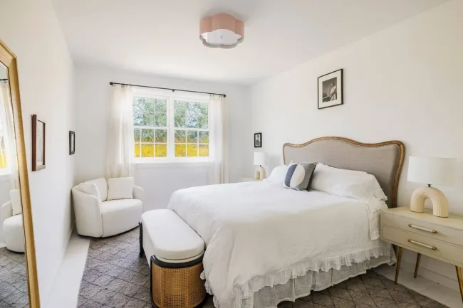 Cozy bedroom with white bed, beige headboard, round chair, bedside tables, and large window.