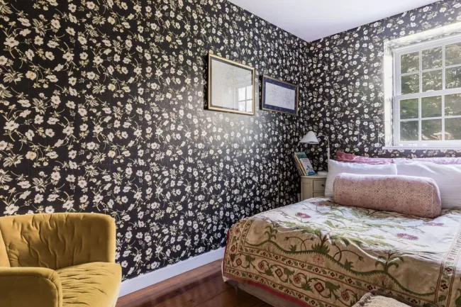 Bedroom with floral wallpaper, yellow chair, bed with patterned bedspread, framed art, and a window.