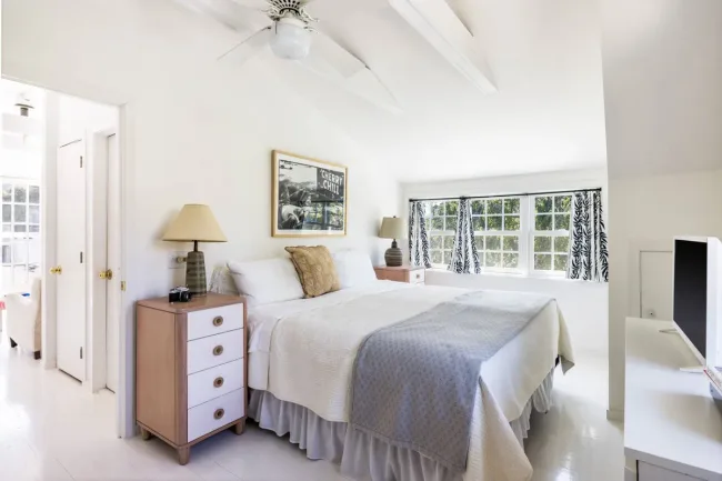 A bright bedroom with a large bed, white bedding, a lamp on a nightstand, large windows, and a "Cherry Chili" poster.