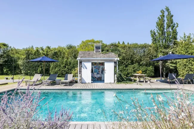 Outdoor swimming pool with loungers, a small pool house, and blue umbrellas surrounded by greenery.