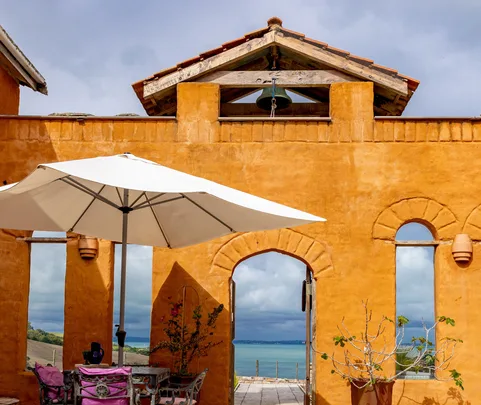 Orange stucco building with archway and patio, white umbrella, pink-cushioned chairs, overlooking a blue sea.