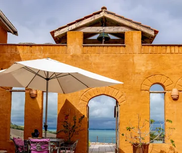 Orange stucco building with archway and patio, white umbrella, pink-cushioned chairs, overlooking a blue sea.