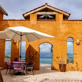 Orange stucco building with archway and patio, white umbrella, pink-cushioned chairs, overlooking a blue sea.