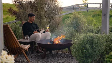 A man drinking a glass of wine with a cat on his lap and a fire burner in front of him.