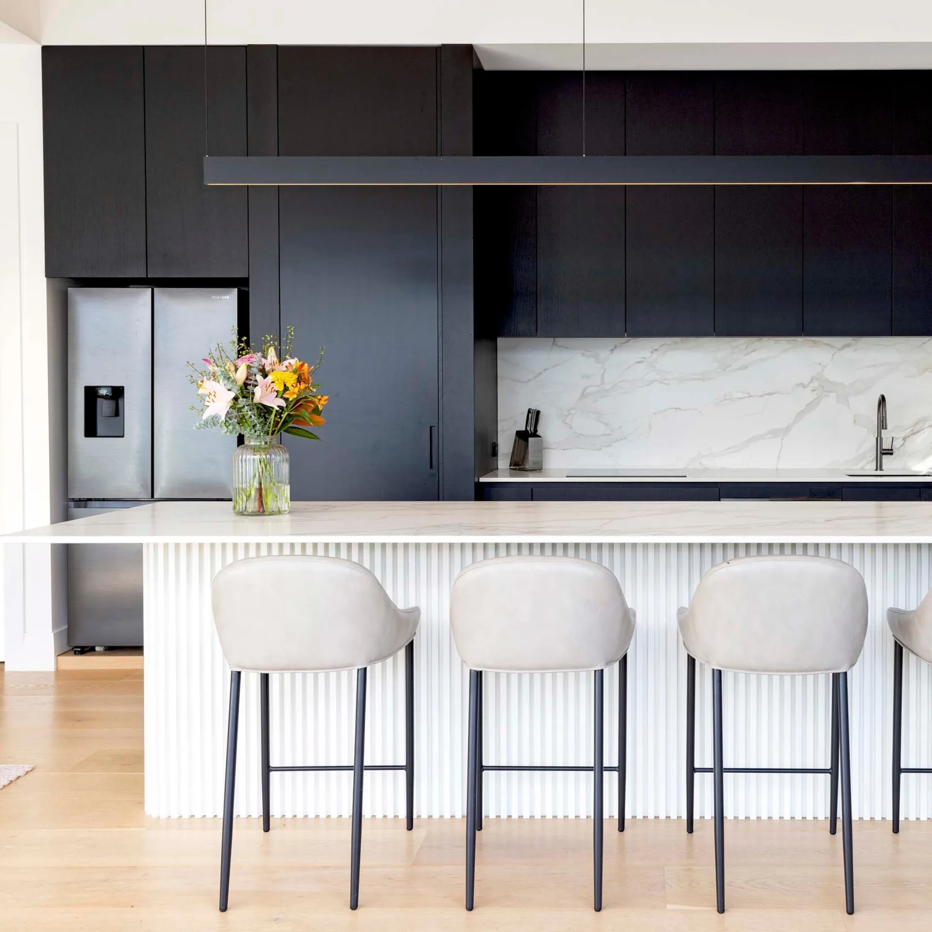 Modern kitchen with black cabinets, marble backsplash, and four stools at a white island. Vase of flowers on the counter.