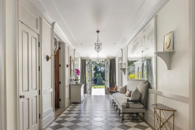 Elegant hallway with checkered floor, plush grey bench, chandeliers, and wall art leading to a garden view.