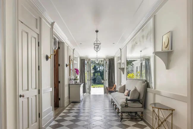 Elegant hallway with checkered floor, plush grey bench, chandeliers, and wall art leading to a garden view.