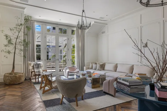 Modern living room with a large beige sectional sofa, tables, books, and tall windows letting in natural light.