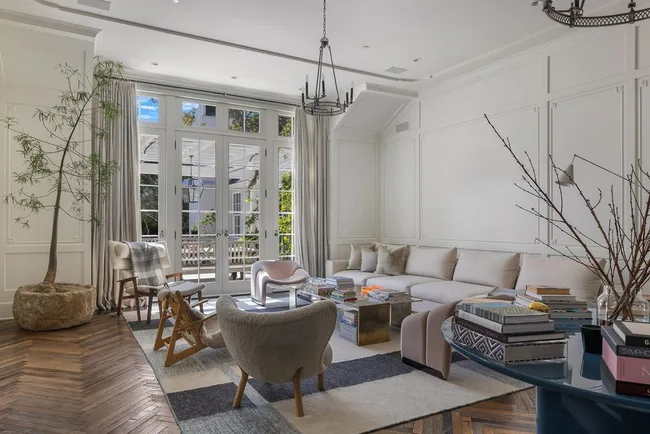 Modern living room with a large beige sectional sofa, tables, books, and tall windows letting in natural light.