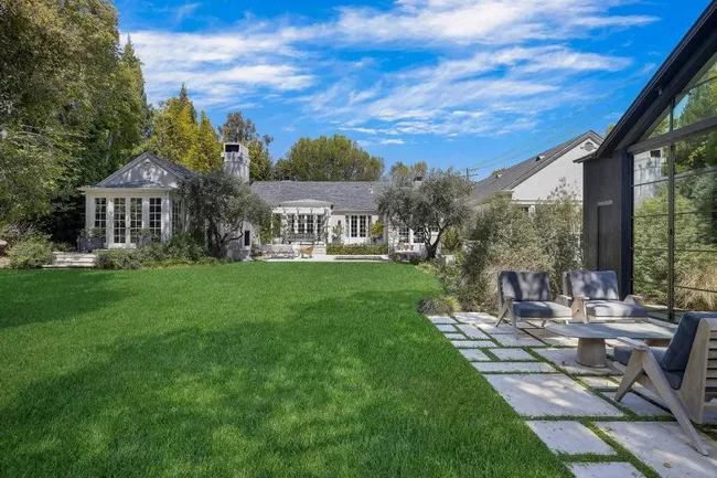 A lush green lawn in front of a large house with tall trees, outdoor seating, and a clear blue sky above.