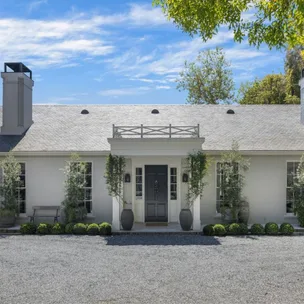 Single-story white brick house with a dark front door, potted plants, and clear blue sky background.