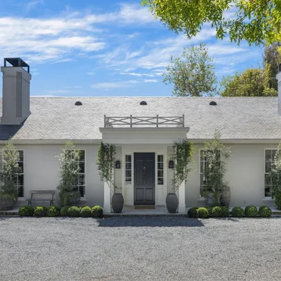 Single-story white brick house with a dark front door, potted plants, and clear blue sky background.