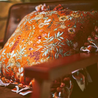 Ornate orange pillow with floral embroidery on a brown chair, casting a cozy, vintage ambiance.