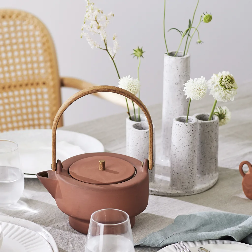 Ceramic teapot with wooden handle on a set table with white flowers in vases and dining ware.