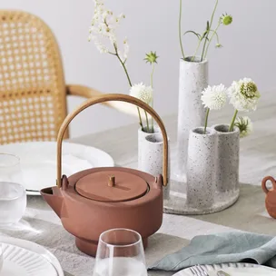 Ceramic teapot with wooden handle on a set table with white flowers in vases and dining ware.