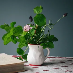 A white jug filled with various green leaves and flowers next to an open book on a patterned cloth.