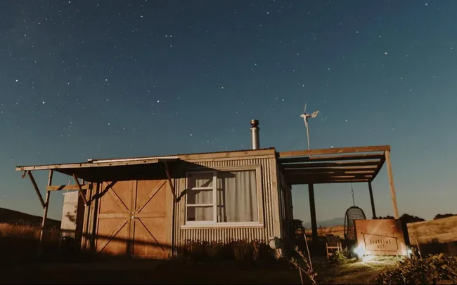 Small rustic hut with night sky and stars, wooden patio, and a swing in rural setting.