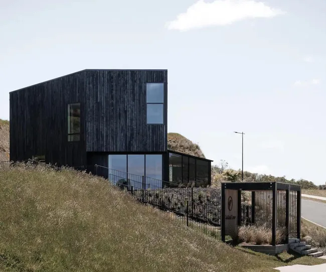 Modern black house with large windows on a sloped grassy hill, with a road and clear sky in the background.