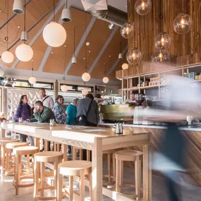 Inside a modern cafe with wooden decor, hanging lights, and people ordering at the counter.