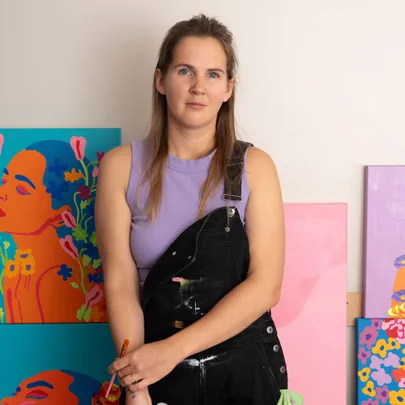 Young woman in overalls holding a paintbrush, stands by colorful paintings with floral motifs in a studio.