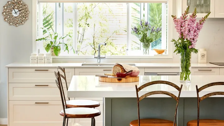Bright kitchen with large window, bar stools, flowers, bread on the island, and a small dog on the hardwood floor.