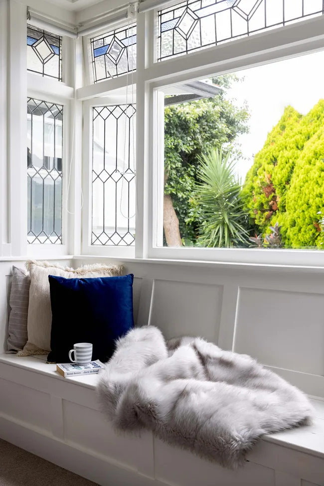Cozy nook with plush pillows and a fur throw by a window, surrounded by greenery and stained glass details.