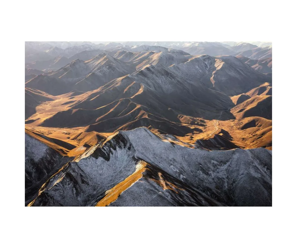 Aerial view of snow-dusted mountain range with warm sunlight casting shadows on the rugged terrain.