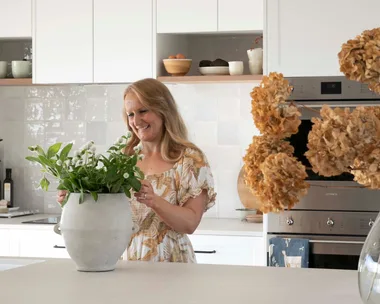 A woman in a kitchen arranging flowers in a vase on a countertop.
