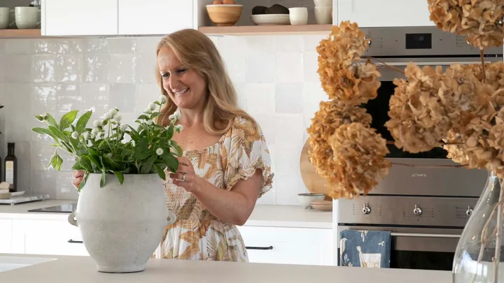 A woman in a kitchen arranging flowers in a vase on a countertop.