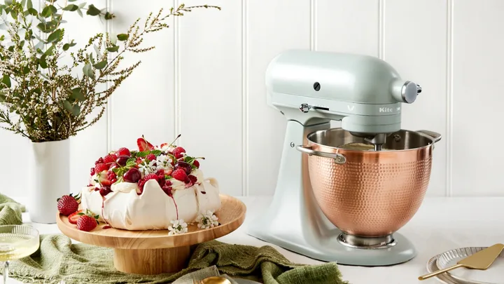 Mixer with copper bowl next to berry-topped cake on wooden stand, white flowers, green towel, and plates.