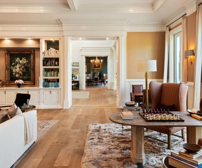 Elegant living room with plush rug, chessboard on table, bookcase, artwork, and view into dining area with chandelier.