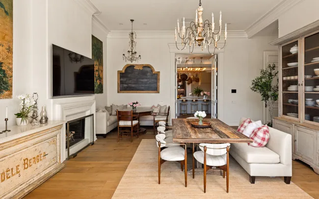 Elegant dining room with wood table, white chairs, chandeliers, fireplace, and decorative art on walls.