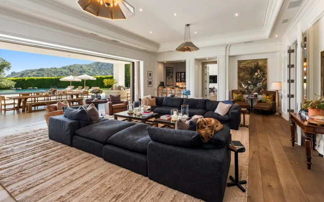 Luxurious living room with dark sofas, open to a patio with dining table, pool, and scenic view in the background.